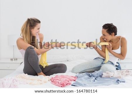 Two pretty female friends jokingly fighting over clothes on bed at home