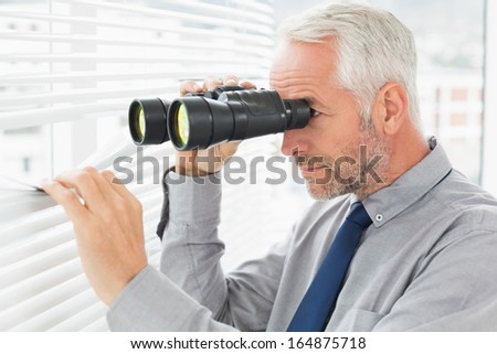 Side view of a serious mature businessman peeking with binoculars through blinds in the office