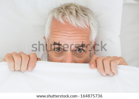 Close-up portrait of a mature shocked man covering face with sheet in bed at home