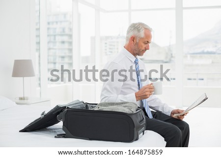 Side view of a mature businessman with coffee cup reading newspaper by luggage at a hotel room