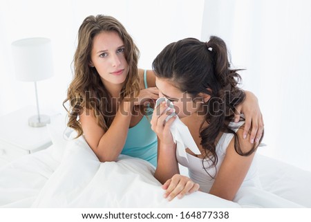 Young woman consoling a crying female friend while sitting on bed at home