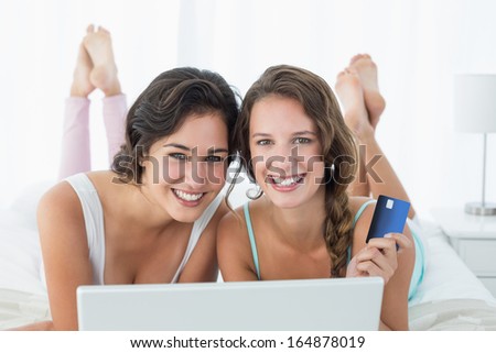 Portrait of happy relaxed young female friends doing online shopping through laptop and credit card in bed