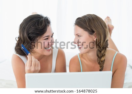 Happy relaxed young female friends doing online shopping through laptop and credit card in bed