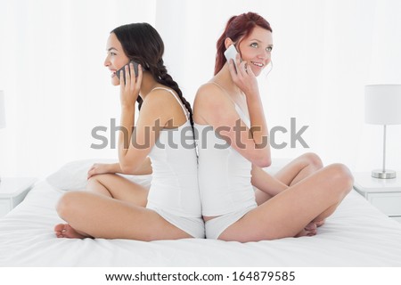 Side view of two happy young female friends using mobile phone while sitting back to back on bed at home
