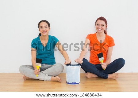 Portrait of two smiling female friends with brushes and paint can sitting in a new house