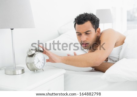 Sleepy young man in bed extending hand to alarm clock