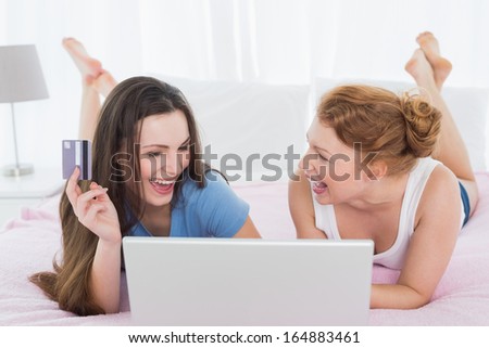 Cheerful relaxed young female friends  doing online shopping through laptop and credit card in bed