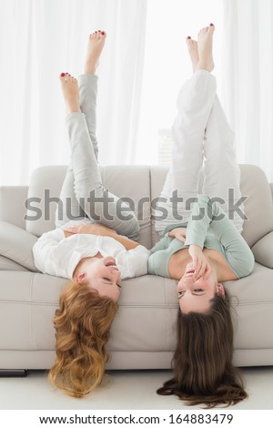 Two smiling young female friends lying on sofa with legs in the air in living room at home