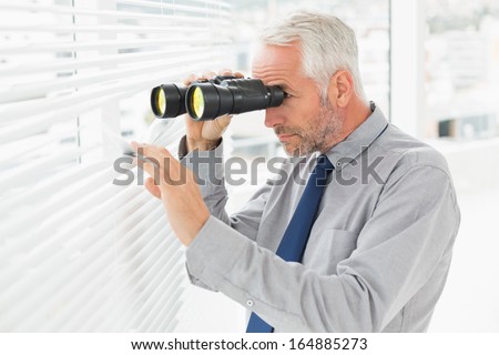 Side view of a serious mature businessman peeking with binoculars through blinds in the office