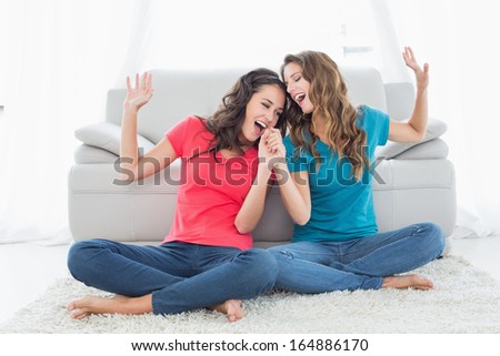 Two Cheerful Young Female Friends Singing In Front Of Sofa In The Living Room At Home