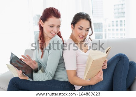 Side view of two relaxed young female friends with digital tablet and book on sofa at home