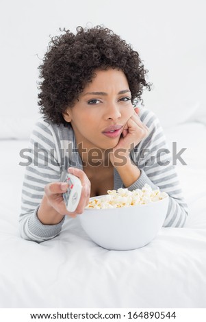 Portrait of a young woman with remote control and popcorn bowl lying in bed at home