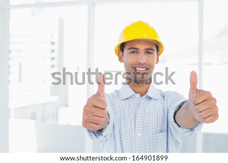 Portrait of a smiling handyman in yellow hard hat gesturing thumbs up in a bright office