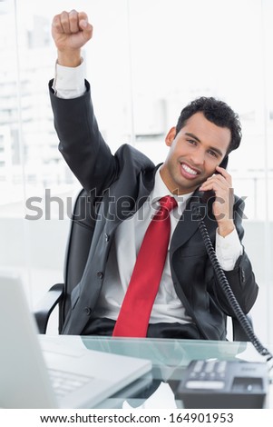Elegant businessman cheering while on call in front of laptop at office desk