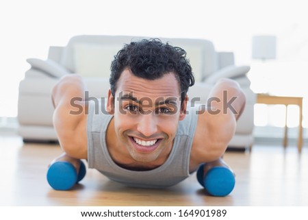 Portrait of a smiling young man with dumbbells doing push ups in the living room at house