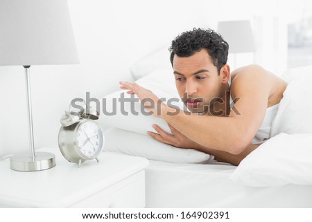 Sleepy young man looking at the alarm clock in bed at home
