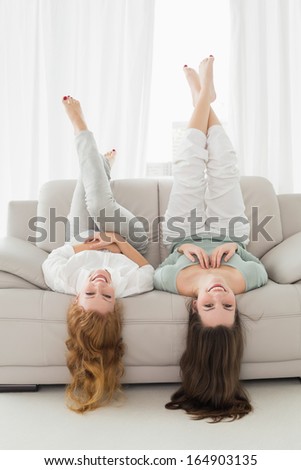 Portrait of two smiling young female friends lying on sofa with legs in the air in living room at home