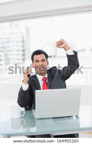 Elegant businessman cheering with clenched fists in front of laptop at office desk