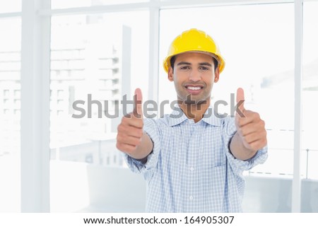 Portrait of a smiling handyman in yellow hard hat gesturing thumbs up in a bright office