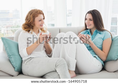 Two happy young female friends with coffee cups conversing in the living room at home
