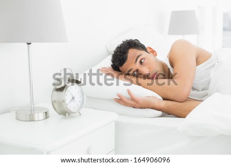 Sleepy young man looking at the alarm clock in bed at home
