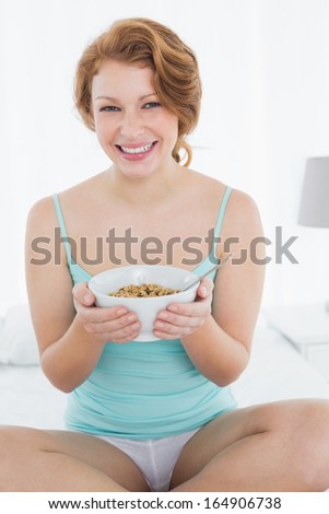 Portrait of a smiling young female with a bowl of cereal sitting on bed at home