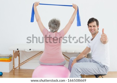 Physical therapist gesturing thumbs up besides senior woman on yoga ball