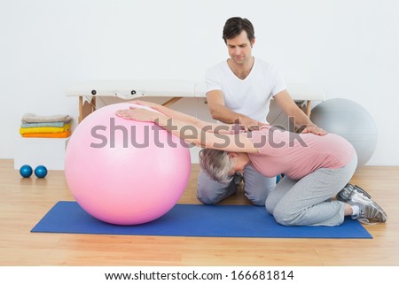 Side view of a physical therapist assisting senior woman with yoga ball in the gym at hospital