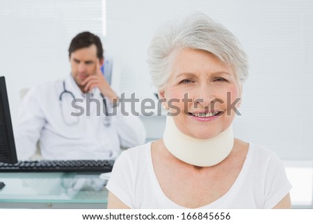 Senior patient in surgical collar with male doctor sitting at desk in background at medical office
