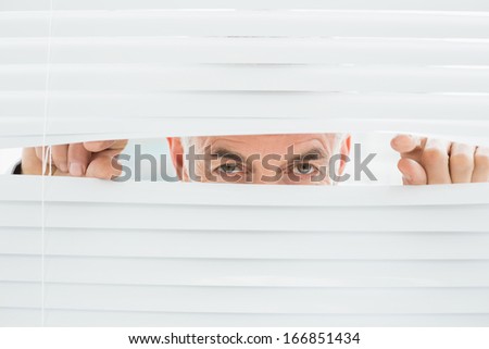 Close-up portrait of a serious mature businessman peeking through blinds in the office