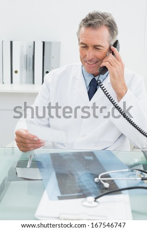 Smiling male doctor using telephone while looking at a note in the medical office