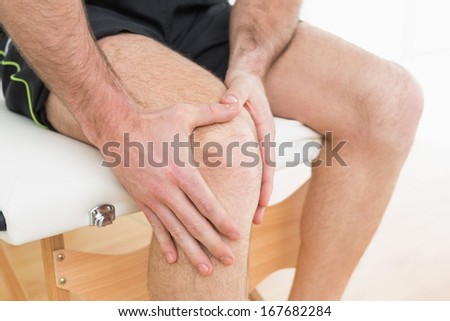 Close-up mid section of a young man with his hands on a painful knee while sitting on examination table