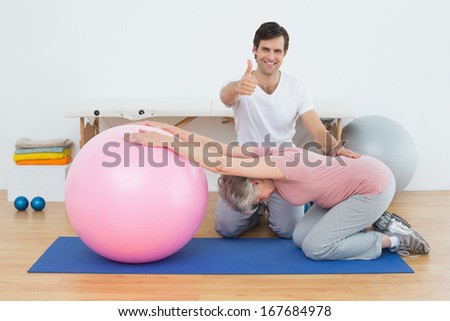 Portrait of a physical therapist gesturing thumbs up by senior woman with yoga ball in the gym at hospital