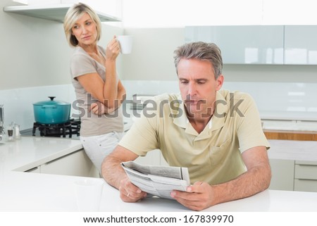 Man reading newspaper while woman standing in background at the kitchen