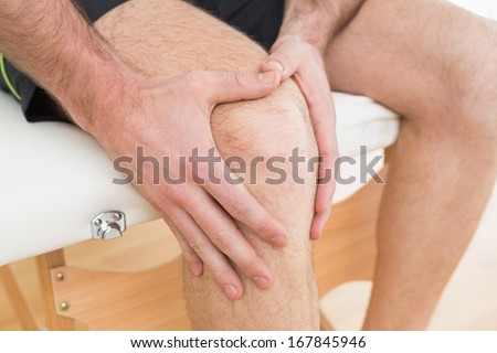 Close-up mid section of a young man with his hands on a painful knee while sitting on examination table