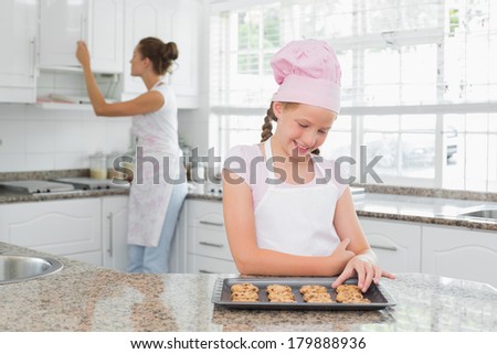 Cute young girl looking at freshly prepared cookies with mother in the background at kitchen