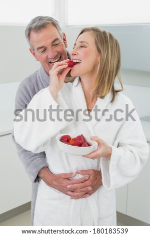 Smiling man embracing woman from behind as she eats strawberry in the kitchen at home