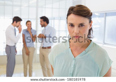 Colleagues gossiping with sad young businesswoman in foreground at a bright office