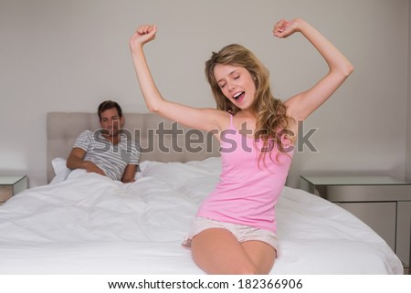 Young woman yawning with man in background on bed at home