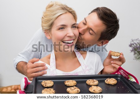 Man kissing woman\'s cheek as she holds freshly baked cookies in the kitchen at home