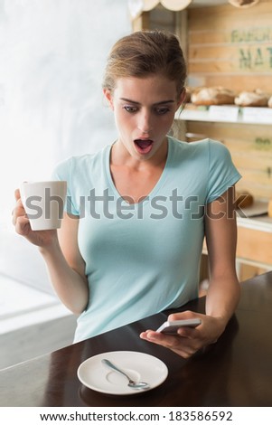 Shocked young woman with coffee cup reading text message at counter in the coffee shop