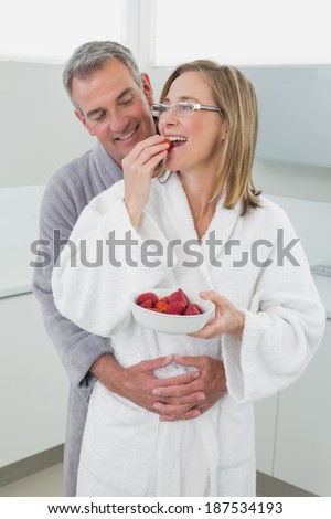 Smiling man embracing woman from behind as she eats strawberry in the kitchen at home