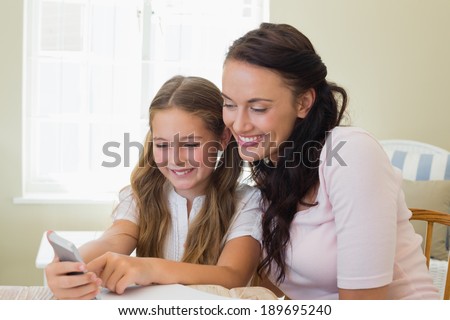 Happy mother and daughter using cell phone together at table in house