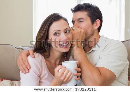 Young man whispering secret into a happy young womans ear in the living room at home