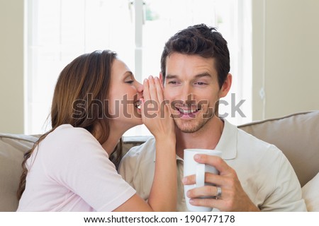 Young woman whispering secret into a happy young mans ear in the living room at home
