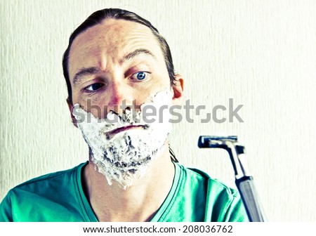 Portrait of bearded young man with foam on face afraid to shaving by razor blade. Image with vintage filter