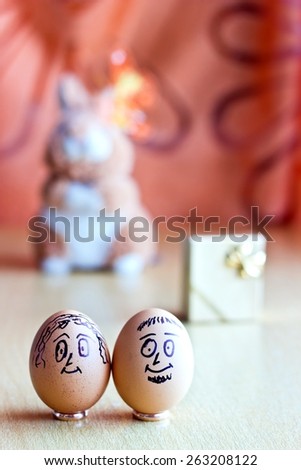Painted easter eggs with man and woman smiling faces. Eggs on wedding rings. Blurred background with golden gift box and easter bunny. Conceptual funny image. Focus on eggs