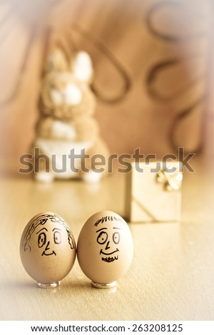 Painted easter eggs with man and woman smiling faces. Eggs on wedding rings. Blurred background with golden gift box and easter bunny. Conceptual funny image. Focus on eggs. Vintage style toned image