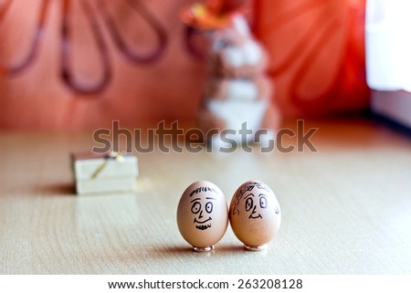 Painted easter eggs with man and woman smiling faces. Eggs on wedding rings. Blurred background with golden gift box and easter bunny. Conceptual funny image. Focus on eggs