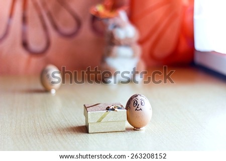 Painted easter eggs with man and woman smiling faces. Eggs on wedding rings. Blurred background with man egg and easter bunny. Conceptual funny image. Focus on woman egg and gift